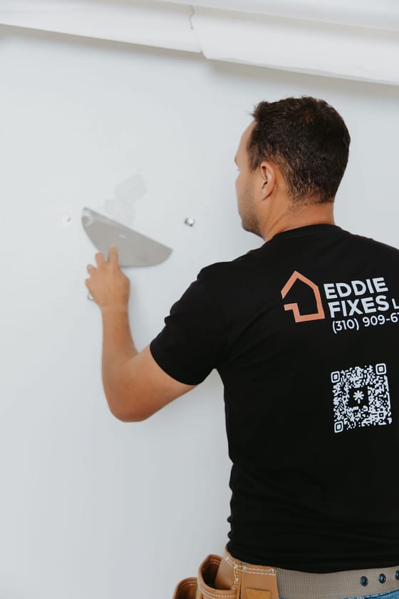 Eddie patching a drywall hole, Eddie Fixes LA shirt seen from behind